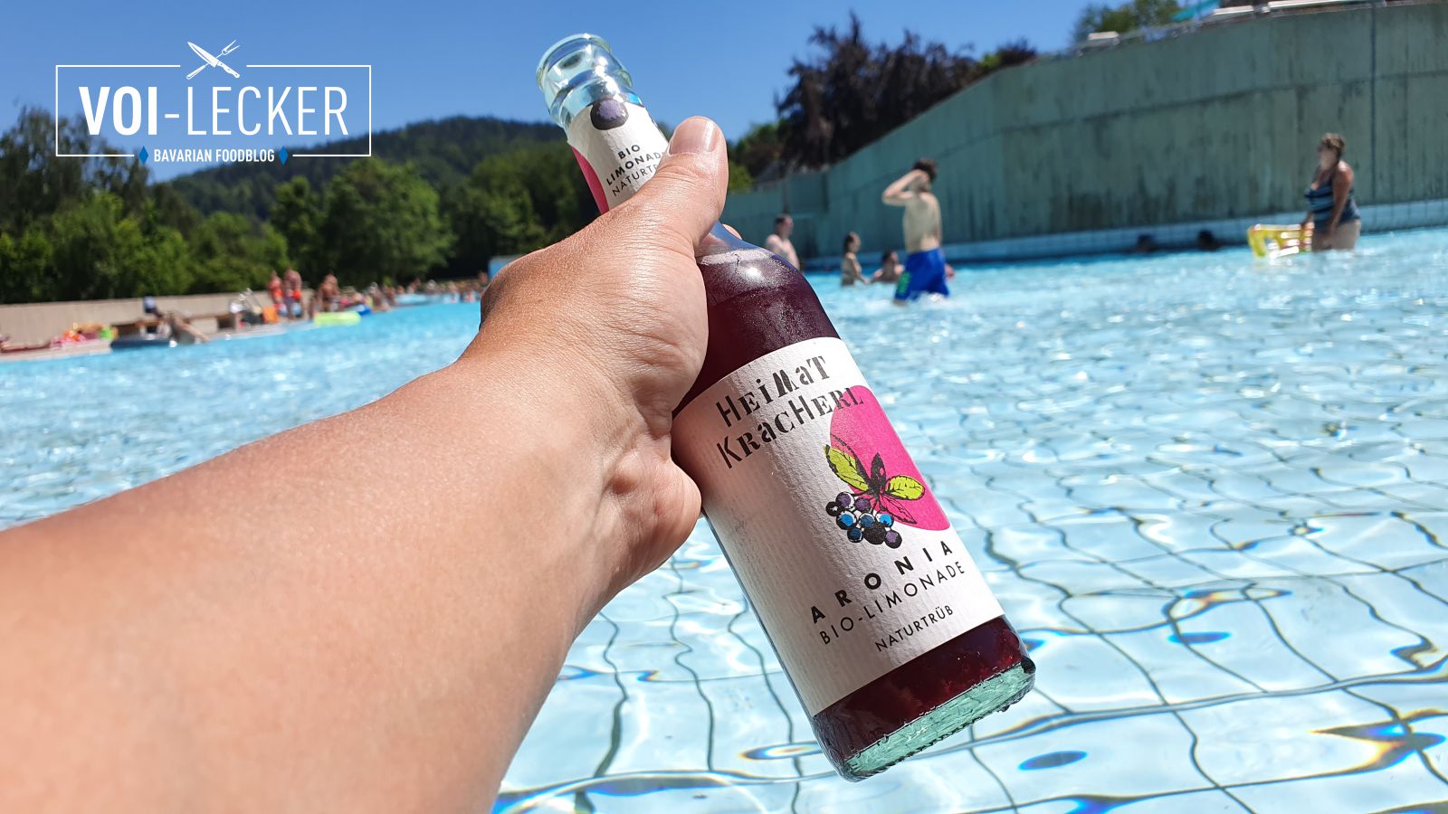 Eine Flasche Heimat Kracherl in der Hand vor einem Freibad